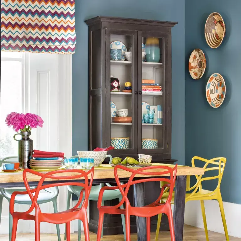 Salle à manger bleue avec des stores à motifs, des chaises et des décorations murales colorées.
