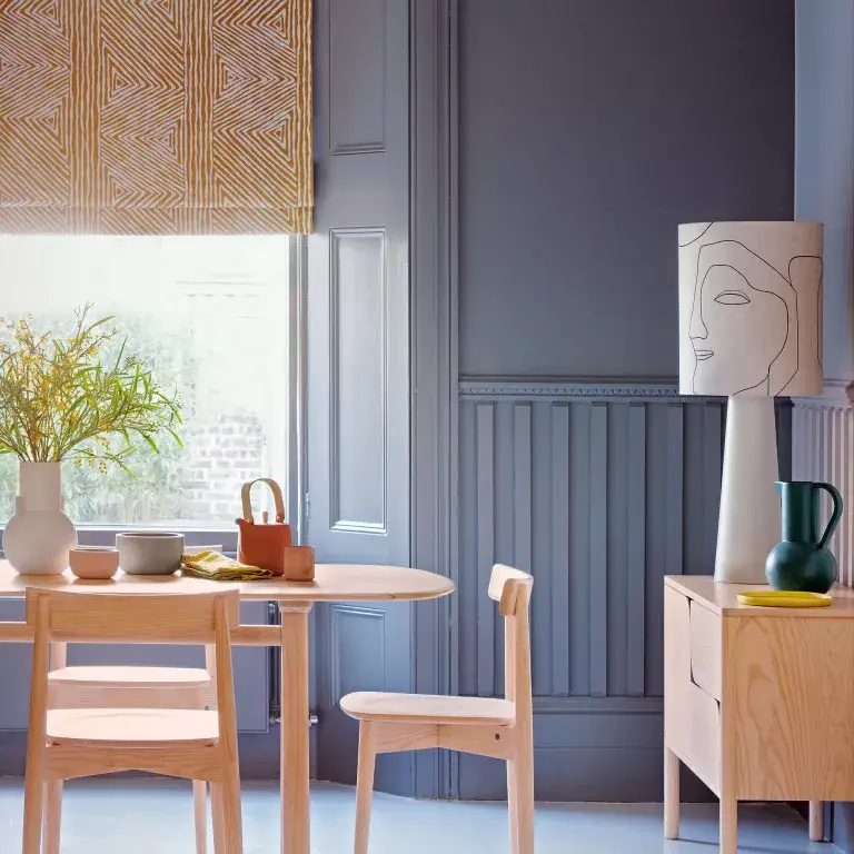 Salle à manger grise avec des meubles en bois clair, une lampe surdimensionnée sur le buffet et un store à motifs sur la fenêtre